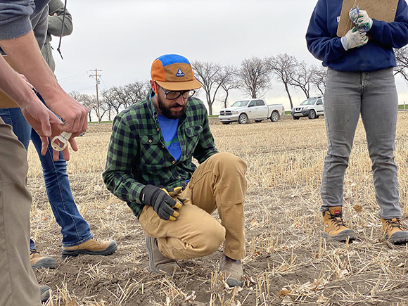 Mike Gretzinger kneels between three students, explaining how to properly hand-dig a seed row to check correct seeding depth and rate.